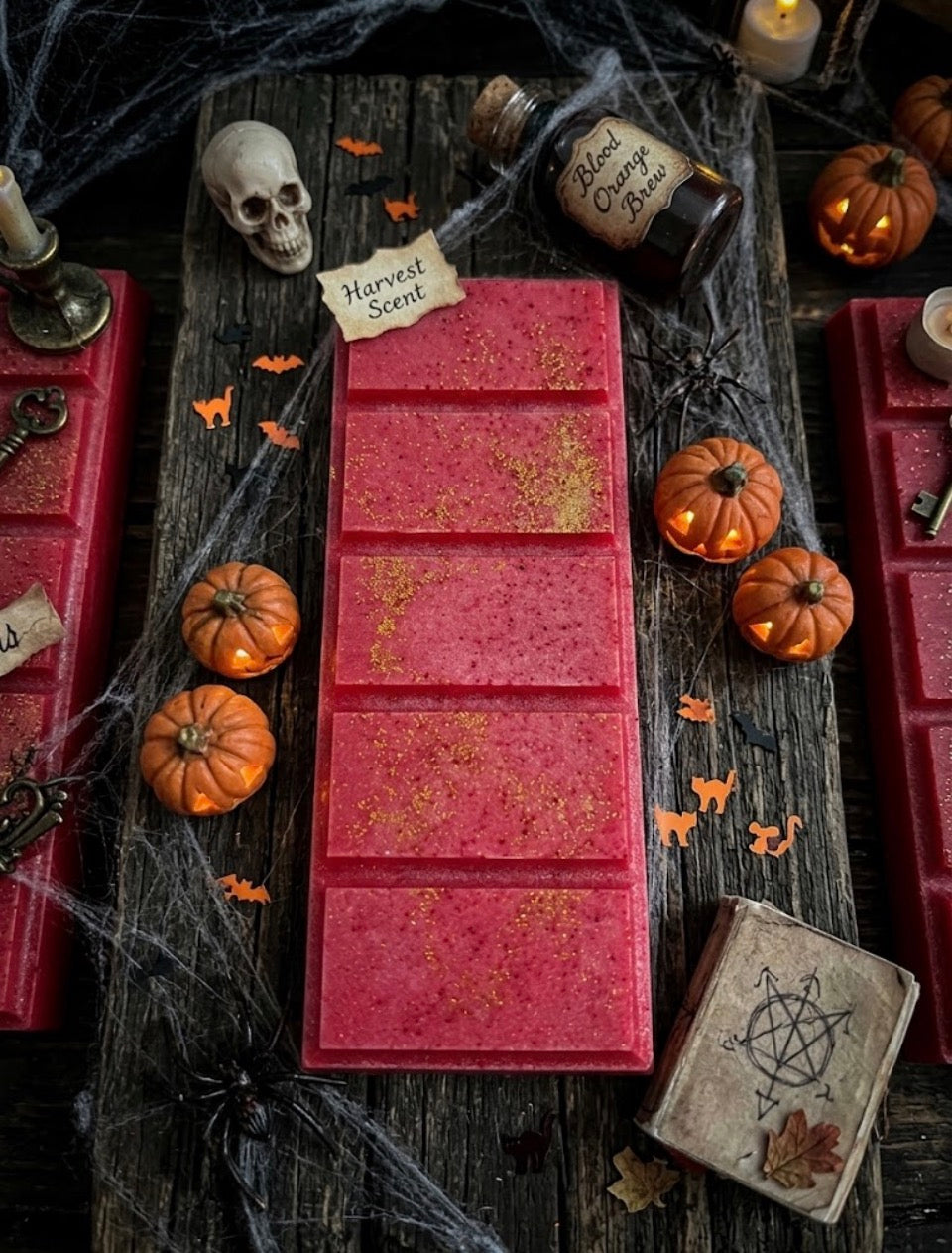 A rectangular pink wax melt bar with gold glitter flakes, displayed on a rustic dark wooden table surrounded by Halloween decorations including small pumpkins, a skull, spiderwebs, and a vintage-style potion bottle.