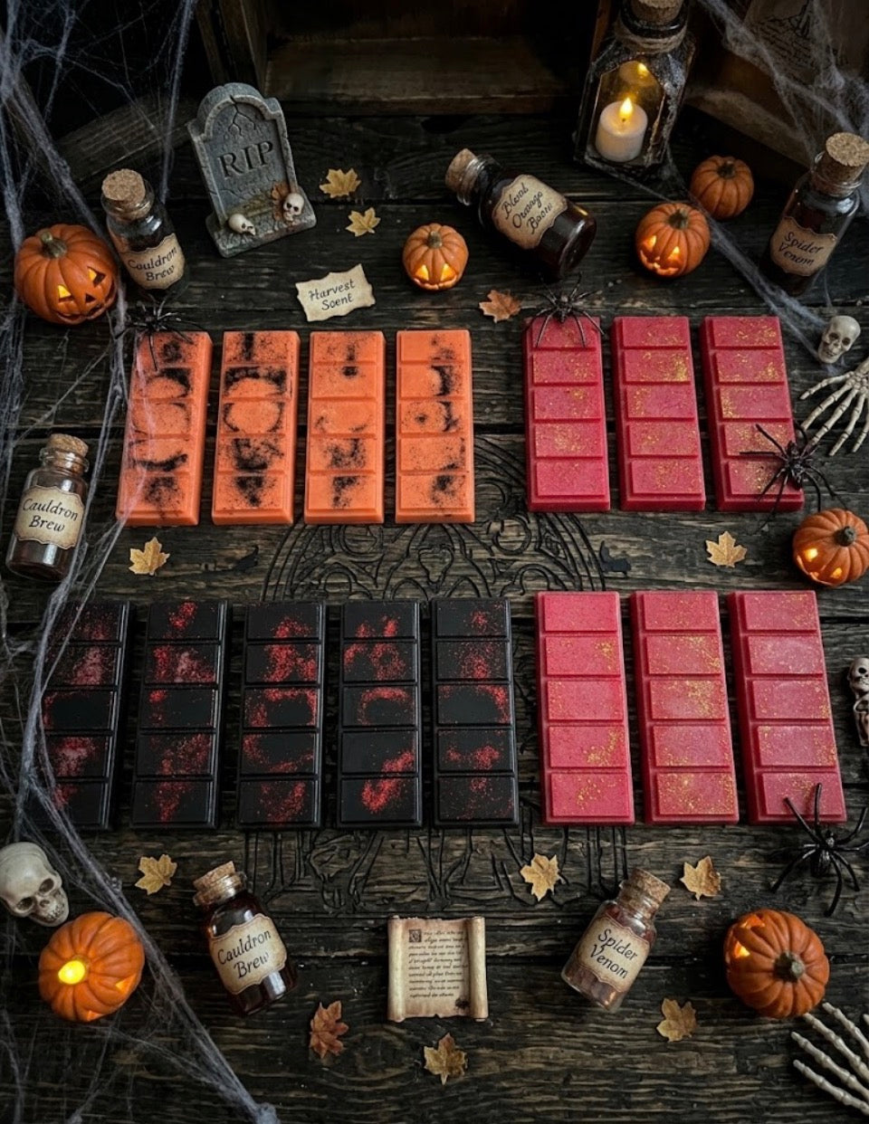 Halloween-themed Wax Melt bar set arranged on a dark wooden table with spooky props including pumpkins, cobwebs, bottles, candles, skulls, and a tombstone. The bars are grouped by color and pattern: orange with black splatter, black with red splatter, and solid red with gold leaf details.