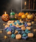 Assorted Halloween shaped wax melts in orange, blue and purple scattered on a rustic wooden surface with jack-o-lanterns, pumpkins, fairy lights and a cauldron essence bottle.