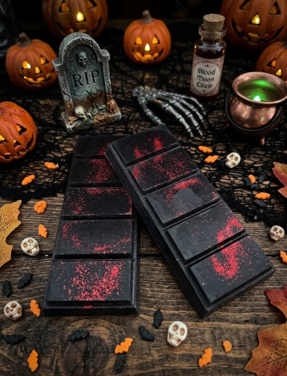 Two Halloween-themed Wax Melts with a dark, blood-splattered finish, displayed on a rustic wooden table surrounded by spooky props including jack-o'-lanterns, a small tombstone, a skeleton hand, and a bottle of blood potion.