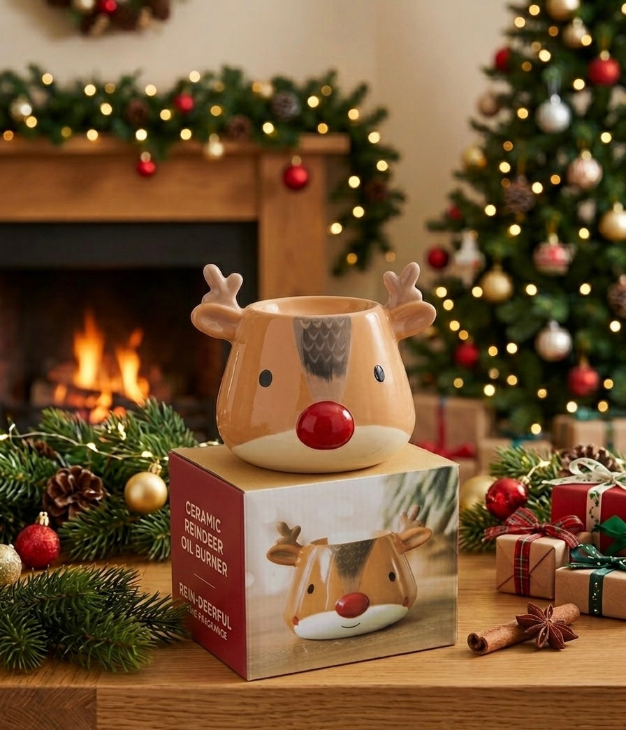 A ceramic reindeer-shaped candle holder with a red nose sits on top of its retail box on a wooden table, surrounded by Christmas greenery, ornaments, a lit fireplace, and wrapped gifts in the background.
