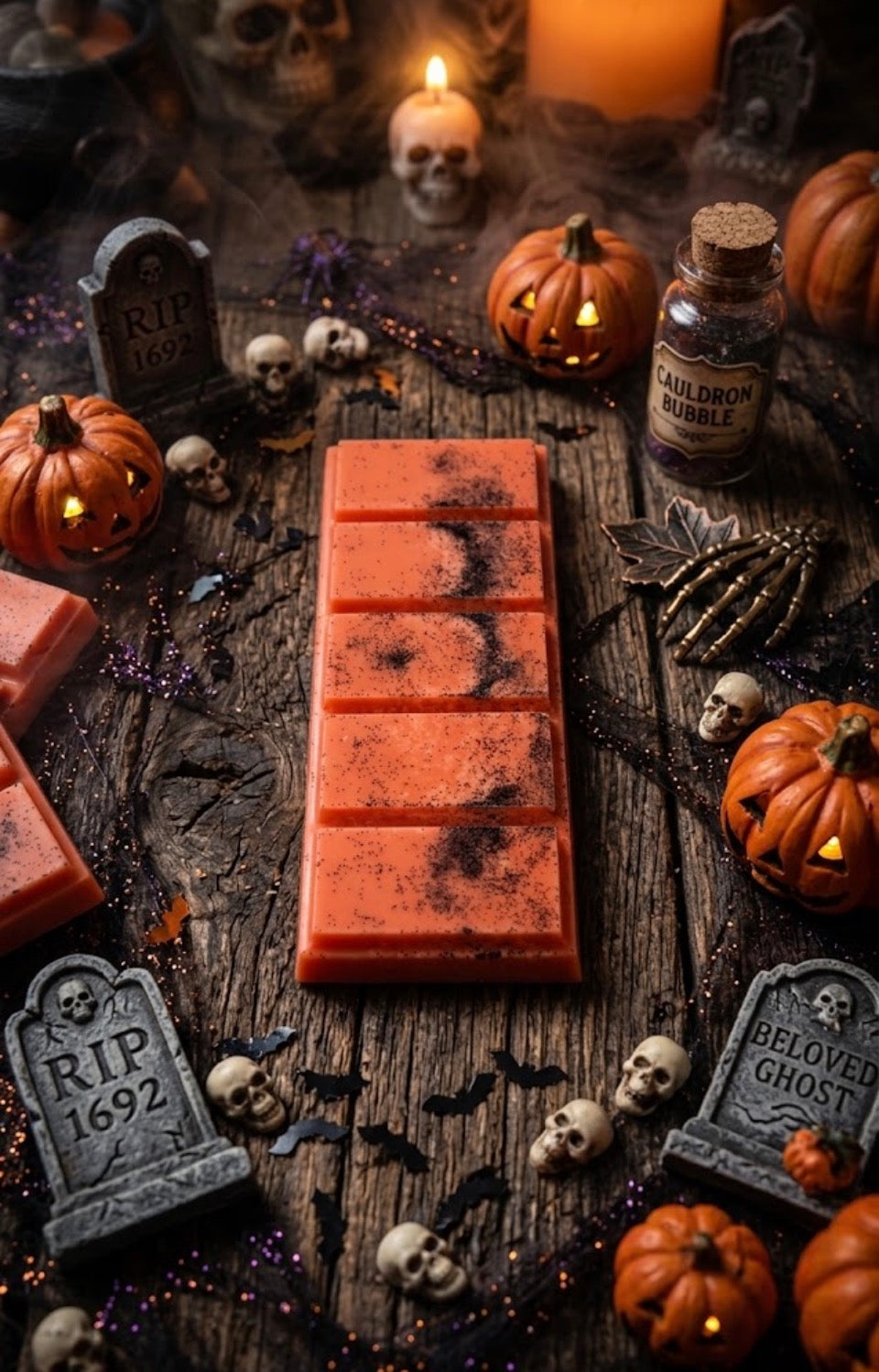 An orange Halloween-themed Wax Melt bar with black splatter details, displayed on a rustic wooden table surrounded by spooky props including miniature tombstones, skulls, jack-o'-lanterns, and a bottle labeled 'Halloween Bubble'.
