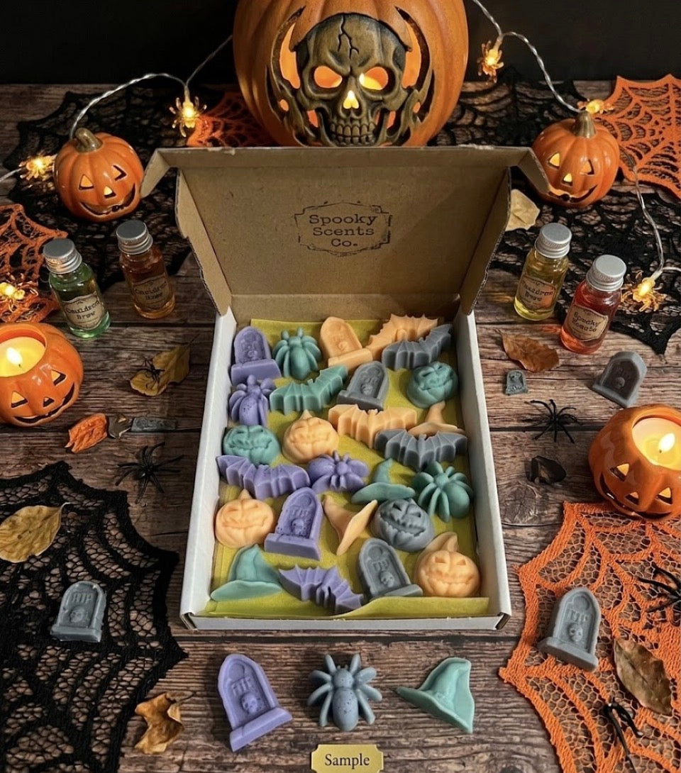 Halloween shaped wax melts in blue, purple and peach arranged in an open gift box on a dark wooden surface surrounded by jack-o-lanterns, cobwebs, spiders and Halloween decorations.