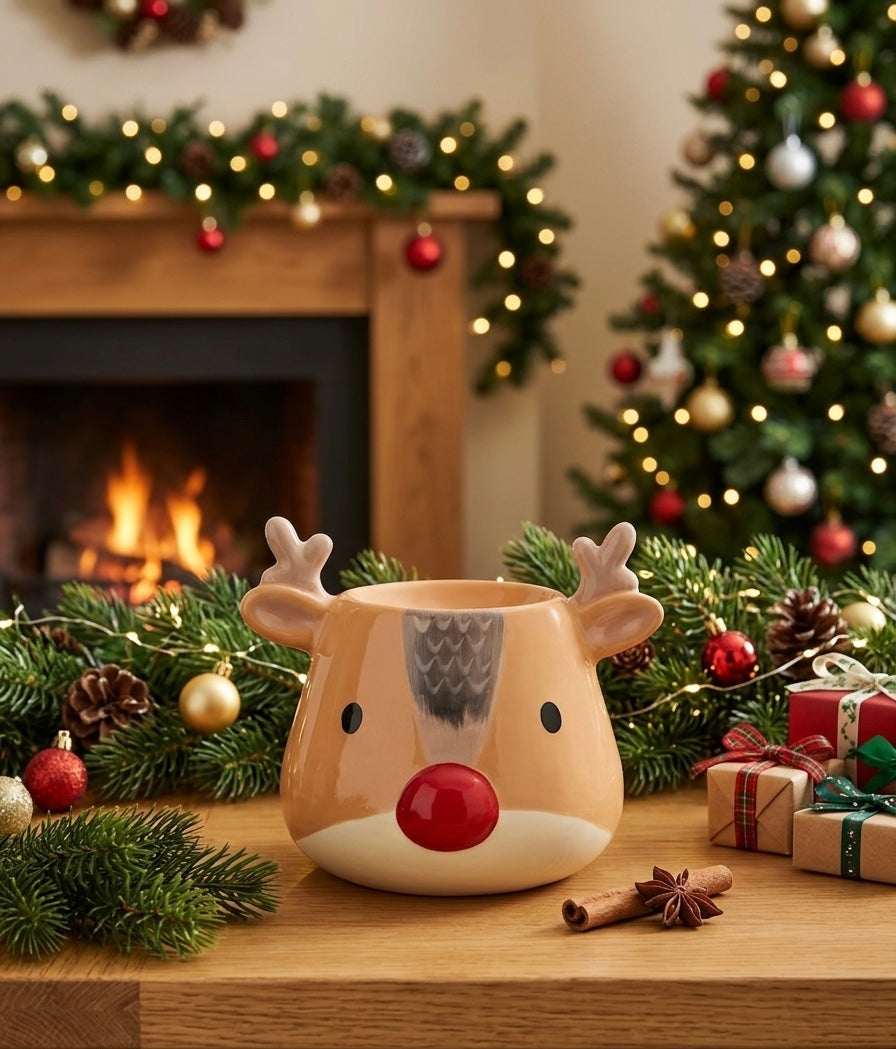 A ceramic oil burner shaped like a reindeer face sits on a wooden table in a cozy holiday setting, with a burning fireplace, garland, Christmas tree, and wrapped gifts in the background.