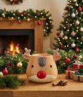 A ceramic oil burner shaped like a reindeer face sits on a wooden table in a cozy holiday setting, with a burning fireplace, garland, Christmas tree, and wrapped gifts in the background.