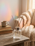 A clear glass reed diffuser bottle with a silver cap and natural wood reeds, placed on a rustic wooden table in a cozy indoor setting with books, a mug, and a knitted blanket in the background.