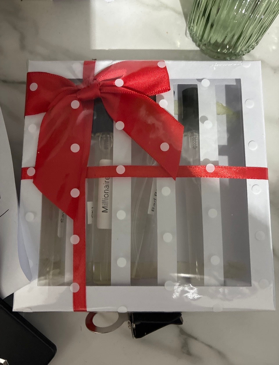 A gift box containing several clear glass perfume bottles, wrapped in white polka dot paper with a large red ribbon bow, placed on a light marble-like surface.